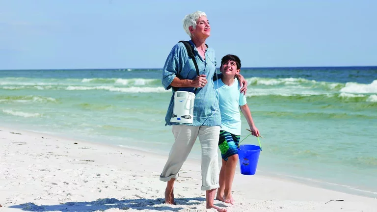 Mulher caminhando na praia com seu neto utilizando um concentrador de oxigênio portátil