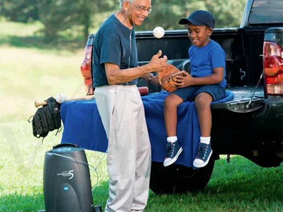 Homem utilizando concentrador transportável de oxigênio conversando com seu neto no parque 