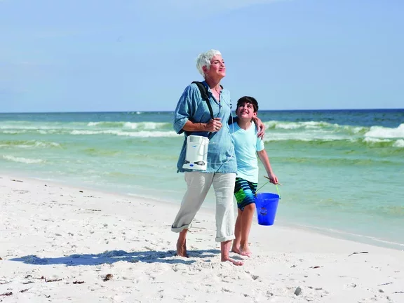 Mulher caminhando na praia com seu neto utilizando um concentrador de oxigênio portátil